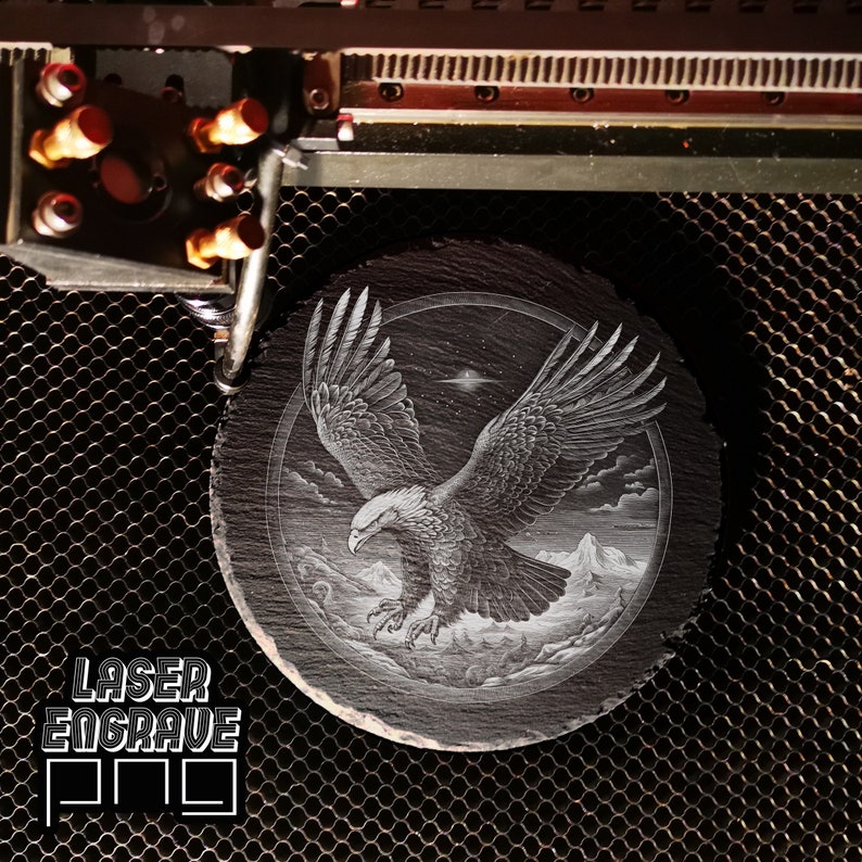 Puede incluir: Un posavasos de pizarra negra con una imagen grabada con l&aacute;ser de un &aacute;guila calva en vuelo. El &aacute;guila vuela sobre una cordillera con nubes en el fondo. La imagen est&aacute; rodeada por un c&iacute;rculo.