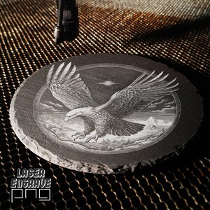 Puede incluir: Un posavasos de pizarra negra con un grabado l&aacute;ser detallado de un &aacute;guila calva en vuelo contra un cielo nocturno estrellado. El &aacute;guila vuela sobre una cordillera con nubes en el fondo.