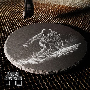 Puede incluir: Un posavasos de pizarra negra con un dise&ntilde;o grabado con l&aacute;ser blanco de un snowboarder en el aire. El snowboarder lleva casco y gafas. El dise&ntilde;o es detallado e intrincado.