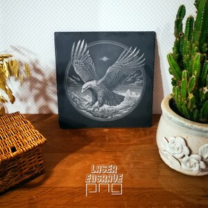 Puede incluir: Un posavasos de pizarra negra con una imagen grabada con l&aacute;ser de un &aacute;guila calva en vuelo. El &aacute;guila est&aacute; volando sobre una cordillera con un cielo estrellado en el fondo. El posavasos tiene el texto "LASER ENGRAVE PRO" impreso en la parte inferior.