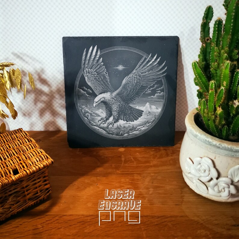 Puede incluir: Un posavasos de pizarra negra con una imagen grabada con l&aacute;ser de un &aacute;guila calva en vuelo. El &aacute;guila est&aacute; volando sobre una cordillera con un cielo estrellado en el fondo. El posavasos tiene el texto "LASER ENGRAVE PRO" impreso en la parte inferior.