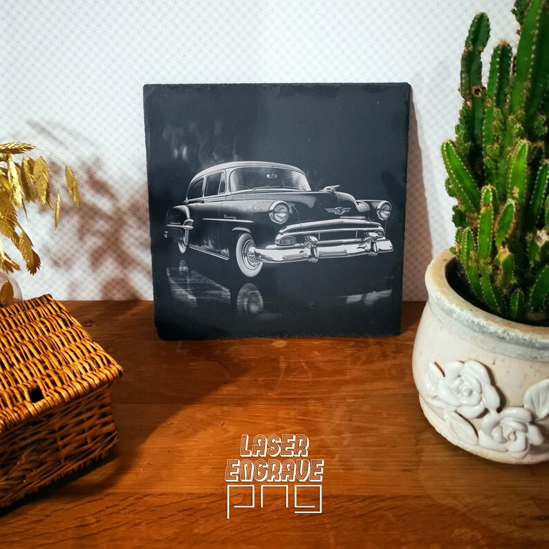 May include: Black and white laser engraved image of a vintage car on a black slate tile. The car is a 1950s Chevrolet with a chrome bumper and grill. The image is set against a dark background.