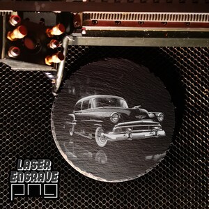 May include: A black and white laser engraved image of a vintage car on a round black slate coaster.