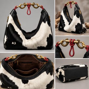 May include: A hobo-style handbag featuring a black and white cow-print pattern with black leather trim. The bag includes a brass-toned metal clasp handle with red leather accents, a zippered top opening, and a brown interior lining with pockets.