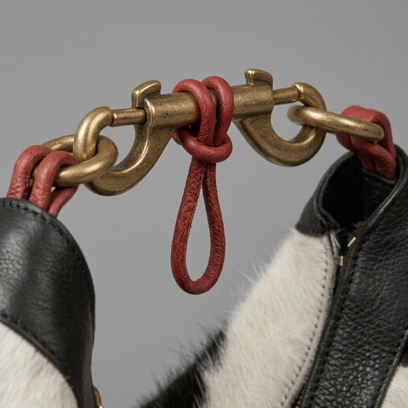 May include: A handbag handle attachment featuring a brass-toned metal swivel clasp connected to black and white textured leather sections by red leather loops. The hardware has a weathered finish, and the red leather is knotted around the metal bar.