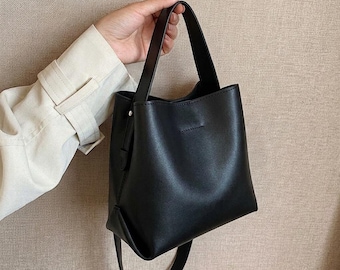 May include: A solid black handbag with a smooth, matte finish, featuring a structured bucket-style silhouette, a single top handle, and a long, thin shoulder strap that hangs down from the base of the bag, creating a clean and simple aesthetic.