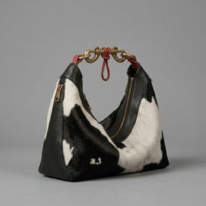 May include: A hobo-style handbag featuring a black and white cowhide pattern with smooth black leather side panels. The bag includes a gold-tone metal clasp handle with red leather accents and a vertical zipper closure on the front exterior.