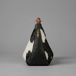 May include: A pyramid-shaped handbag featuring a black and white cowhide pattern on the sides and a solid black leather front panel. The bag includes a gold-toned vertical zipper closure and a short, red leather top handle with gold hardware.