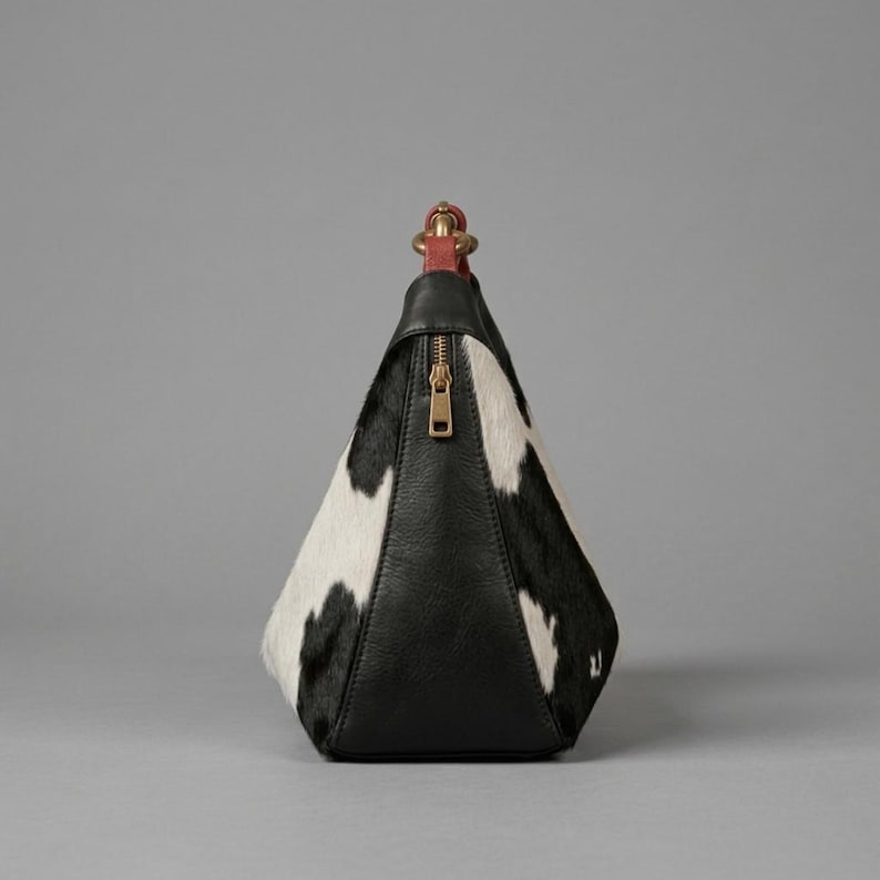 May include: A pyramid-shaped handbag featuring a black and white cowhide pattern on the sides and a solid black leather front panel. The bag includes a gold-toned vertical zipper closure and a short, red leather top handle with gold hardware.