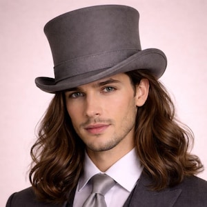 Puede incluir: Un sombrero de copa gris con ala ancha y una banda alrededor de la corona. El sombrero se usa con un traje gris, camisa blanca y corbata gris. La persona tiene el pelo largo y castaño.