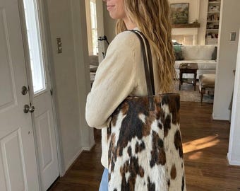 May include: A tote bag featuring a cowhide pattern with patches of brown, black, and white fur. The bag has two long, thin, dark brown leather shoulder straps attached to the top edge, providing a vertical rectangular shape for carrying items.