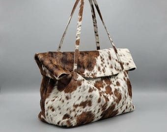 May include: A tote bag crafted from cowhide featuring a brown and white spotted pattern. The bag has a fold-over top design and two thin, matching cowhide shoulder straps. The texture of the material is visible, showing the natural hair of the hide.