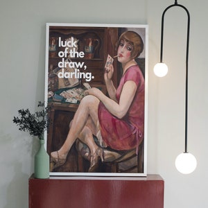 Puede incluir: Una impresión enmarcada de una mujer con un vestido rosa sentada en una mesa con cartas y una copa de vino. El texto "luck of the draw, darling." está impreso en blanco sobre un fondo negro.