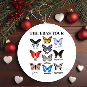 Puede incluir: Un adorno redondo blanco con el texto "THE ERAS TOUR" e imágenes de mariposas que representan cada uno de los álbumes de Taylor Swift. Las mariposas están etiquetadas con los nombres de los álbumes: "Fearless", "Speak Now", "1989", "Red", "reputation", "folklore", "Lover", "Midnights" y "evermore".