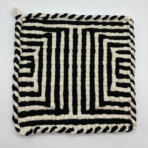 May include: Square black and white woven pot holder with a loop for hanging. The design features alternating vertical black and white stripes, creating a geometric pattern. The edges are finished with a black rope-like trim.