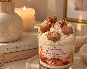 Puede incluir: Una vela en frasco de vidrio transparente con remolinos de cera blanca, decoraciones en miniatura de rollos de canela marrones y un anís estrellado. La etiqueta dice La Belle Lumiere London Cinnamon Bun Scented Candle.