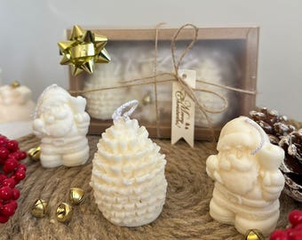 Puede incluir: Velas navideñas de color crema con forma de Papá Noel y piña. Las velas se exhiben con campanas doradas, bayas rojas y una caja de regalo atada con hilo. También hay un lazo dorado y una etiqueta que dice "Merry Christmas".