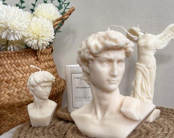 Puede incluir: Velas de busto blanquecinas de esculturas clásicas, incluyendo a David y la Victoria de Samotracia. Las velas están sobre una esterilla de yute, con una cesta de flores blancas en el fondo. También es visible una vela con el texto "GINGERBREAD".