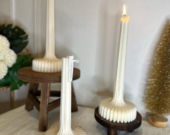 Puede incluir: Tres velas de pilar de color marfil con una base acanalada y tallos altos y delgados. Una vela está encendida, mostrando una pequeña llama. Las velas están sobre una mesa de madera y un pequeño taburete de madera, con un fondo neutro.