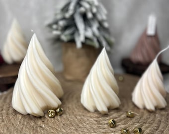 Puede incluir: Velas en forma de árbol arremolinado de color crema con campanas doradas sobre una estera tejida. Un pequeño árbol de Navidad cubierto de nieve está en el fondo. Las velas son una decoración festiva para la temporada navideña.