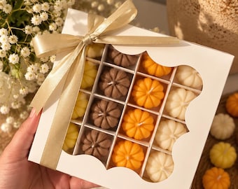 Puede incluir: Una caja de regalo cuadrada blanca con una ventana festoneada que contiene dieciséis pequeñas velas con forma de calabaza en tonos marrón, naranja, amarillo y crema. La caja está atada con un lazo de satén dorado. Hay velas de calabaza sueltas cerca.