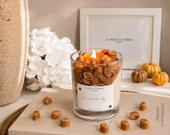 Puede incluir: Una vela en vaso de cristal transparente rellena de cera marrón claro y decorada con numerosos trozos de cera pequeños con forma de grano de café. Una etiqueta blanca frontal dice LA BELLE LUMIERE LONDON, COFFEE BEAN y 100 SOY WAX CANDLE. La vela está encendida.