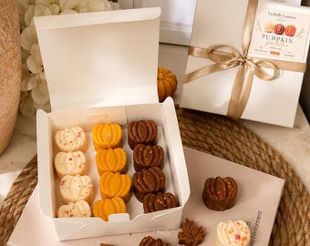 Puede incluir: Una caja de cartón blanca que contiene doce ceras aromáticas con forma de calabaza en colores blanco, naranja y marrón con motas. Otras ceras sueltas con forma de calabaza y hoja están dispuestas sobre un libro titulado Artful Lifestyle.