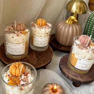 Puede incluir: Cuatro velas de cristal con cera naranja, cubiertas con cera batida blanca, peque&ntilde;as calabazas decorativas y hojas de oto&ntilde;o. Cada etiqueta blanca dice La Belle Lumiere London Pumpkin Spice. Una vela con forma de calabaza encendida est&aacute; cerca.