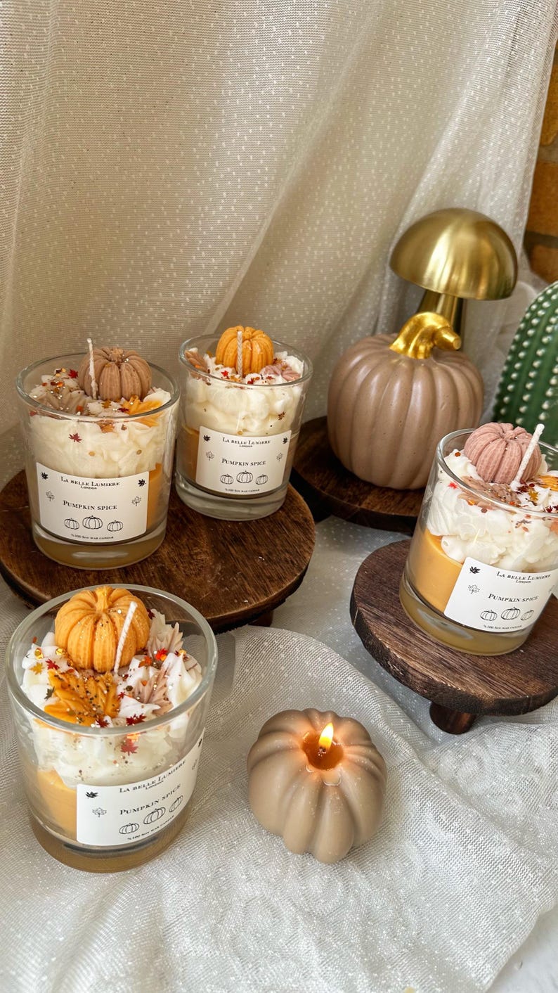 Puede incluir: Cuatro velas de cristal con cera naranja, cubiertas con cera batida blanca, peque&ntilde;as calabazas decorativas y hojas de oto&ntilde;o. Cada etiqueta blanca dice La Belle Lumiere London Pumpkin Spice. Una vela con forma de calabaza encendida est&aacute; cerca.