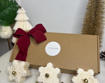 Puede incluir: Un juego de regalo festivo con tres derretidos de cera en forma de copo de nieve, una caja de cartón marrón y un lazo rojo. Los derretidos son blancos con detalles dorados. La caja tiene el texto "LA BELLE LUMIERE LONDON".