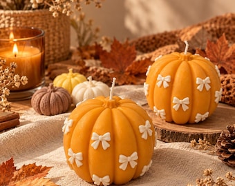 Puede incluir: Dos velas naranjas con forma de calabaza decoradas con pequeños lazos blancos. Las velas presentan una textura acanalada y una mecha central única. Están dispuestas junto a velas más pequeñas con forma de calabaza en amarillo, blanco y marrón.