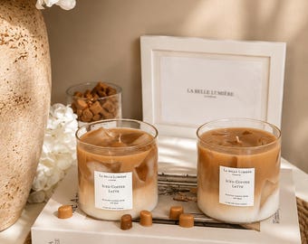 Puede incluir: Dos velas de cristal con un diseño de cera en capas marrón y blanco que recuerda al café helado. Cada vela tiene una etiqueta blanca que dice La Belle Lumiere London Iced Coffee Latte. Pequeños trozos de cera marrón están esparcidos.