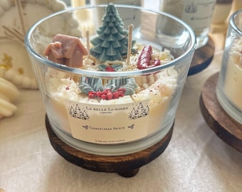 Puede incluir: Un portavelas de cristal transparente con base de madera, relleno de cera blanca y elementos decorativos. La vela presenta una casa en miniatura, un árbol de Navidad y el texto "Christmas Spice".