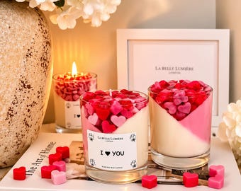 Puede incluir: Dos velas de cristal rellenas de cera color crema y cubiertas con pequeñas piezas de cera en forma de corazón rojas y rosas. La vela frontal tiene una etiqueta blanca con el texto LA BELLE LUMIERE LONDON e I HEART YOU. Corazones de cera rojos y rosas esparcidos.