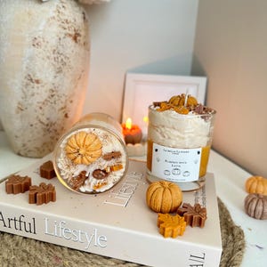 Puede incluir: Dos velas en frasco de vidrio con cera color crema decoradas con peque&ntilde;as calabazas y hojas de cera. Una vela est&aacute; de pie con una etiqueta La Belle Lumiere Pumpkin Spice Latte, la otra est&aacute; acostada sobre un libro titulado Artful Lifestyle.