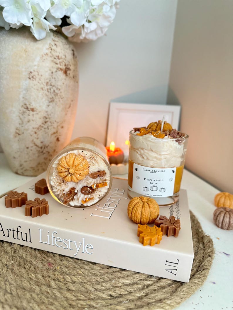 Puede incluir: Dos velas en frasco de vidrio con cera color crema decoradas con peque&ntilde;as calabazas y hojas de cera. Una vela est&aacute; de pie con una etiqueta La Belle Lumiere Pumpkin Spice Latte, la otra est&aacute; acostada sobre un libro titulado Artful Lifestyle.