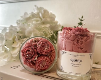 Puede incluir: Una vela en vaso de cristal transparente con cera rosa en forma de ramo de flores, decorada con purpurina dorada. La etiqueta blanca dice La Belle Lumiere London, Pinky Flower Bouquet, 100% Soy Wax Candle. Se ve una segunda vela desde arriba.