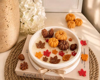 Puede incluir: Un recipiente redondo y blanco para velas decorado con una cinta beige, relleno de cera y cubierto con pequeñas figuras de cera de calabazas, piñas, bellotas y hojas de otoño en tonos naranja, marrón y rojo, sobre un libro titulado Artful Lifestyle.