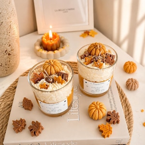 Puede incluir: Dos velas de cristal con cera blanca batida en la parte superior, decoradas con peque&ntilde;as calabazas y hojas de arce de cera. Las etiquetas dicen La Belle Lumiere Pumpkin Spice Latte. Peque&ntilde;as calabazas y hojas de cera descansan sobre un libro.