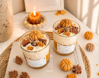 Op de afbeelding: Twee glazen kaarsen met een witte opgeklopte waslaag, versierd met kleine wassen pompoenen en esdoornbladeren. De etiketten vermelden La Belle Lumiere Pumpkin Spice Latte. Kleine wassen pompoenen en blaadjes liggen verspreid op een boek.