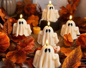 Puede incluir: Cinco velas de color blanco roto con forma de fantasma y ojos pintados en negro, cada una con una mecha encendida en la parte superior. Las velas están dispuestas entre hojas de otoño naranjas y marrones y pequeñas calabazas decorativas.
