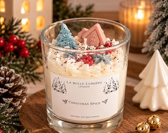 Puede incluir: Una vela en vaso de cristal transparente con cera de soja blanca, decorada con figuras de cera que incluyen un árbol azul, una casa rosa y bayas rojas. La etiqueta dice La Belle Lumière London Christmas Spice Natural Soy Wax Candle.