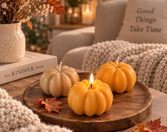 May include: Three pumpkin-shaped candles arranged on a round wooden tray. One candle is lit, displaying a bright flame, while the other two are unlit. The candles are colored in shades of beige and orange, featuring realistic vertical ridges.