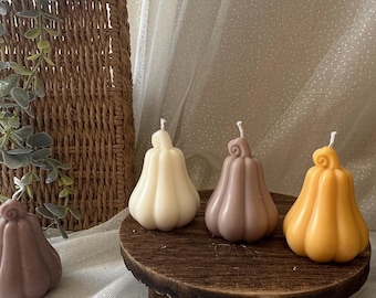 Puede incluir: Velas con forma de calabaza en tonos crema, malva y naranja. Las velas se exhiben en un pequeño soporte redondo de madera. El fondo incluye una cesta tejida y tela drapeada.