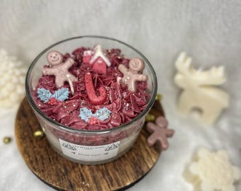 Puede incluir: Una vela de vidrio con una parte superior de color burdeos, decorada con hombres de jengibre, una casa y acebo. La vela se encuentra sobre una base de madera. La etiqueta dice "LA BELLE LUMIERE" y "WINTER SEASON".