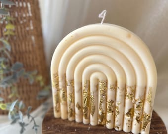 Puede incluir: Una vela en forma de arcoíris de color crema con detalles de pan de oro. La vela está sobre un pequeño soporte redondo de madera. La vela tiene una mecha en la parte superior y no tiene perfume. Es un artículo decorativo.