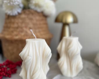 Puede incluir: Dos velas blanquecinas texturizadas con un diseño ondulado. Las velas son cilíndricas y están sobre una superficie blanca. En el fondo hay una cesta de mimbre con flores blancas, junto con un objeto dorado con forma de hongo.