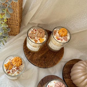 Puede incluir: Tres velas de vidrio cubiertas con cera blanca, figuras de cera en forma de calabaza naranja y beige, y peque&ntilde;os detalles de cera en forma de hojas coloridas. Cada vela tiene una etiqueta blanca con texto. Tambi&eacute;n hay dos figuras de cera de calabaza.