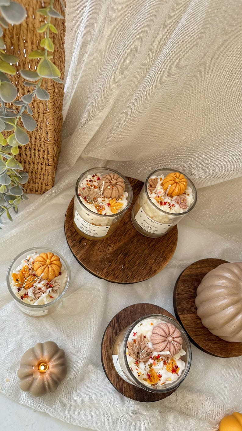 Puede incluir: Tres velas de vidrio cubiertas con cera blanca, figuras de cera en forma de calabaza naranja y beige, y peque&ntilde;os detalles de cera en forma de hojas coloridas. Cada vela tiene una etiqueta blanca con texto. Tambi&eacute;n hay dos figuras de cera de calabaza.