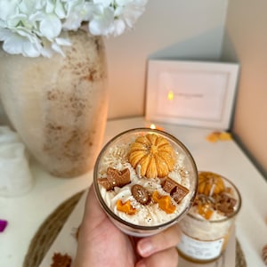 Puede incluir: Un recipiente de vidrio para velas relleno de cera blanca con una calabaza naranja decorativa, hojas de arce marrones y peque&ntilde;as incrustaciones de cera en forma de estrella. La superficie tiene purpurina dorada y textura batida.